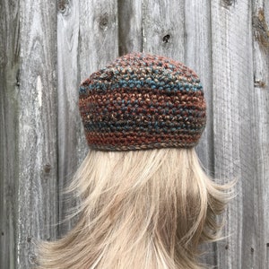 Op de afbeelding: Een gebreide beanie met een bruin, blauw en beige gestreept patroon. De beanie is gemaakt van een getextureerd garen en heeft een slouchy pasvorm.