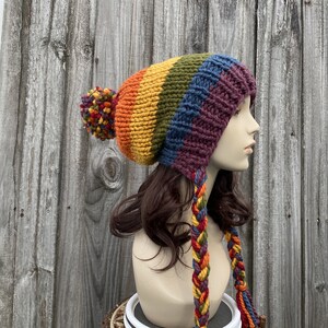 Pu&ograve; includere: Un berretto di lana colorato con un pon pon e lunghi lacci intrecciati. Il berretto &egrave; realizzato in un motivo arcobaleno con sfumature di rosso, arancione, giallo, verde, blu e viola.