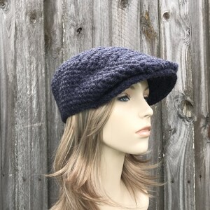 May include: A navy blue newsboy cap made of a textured knit fabric. The cap has a rounded crown and a short, stiff brim. The hat is displayed on a mannequin against a weathered wooden background.
