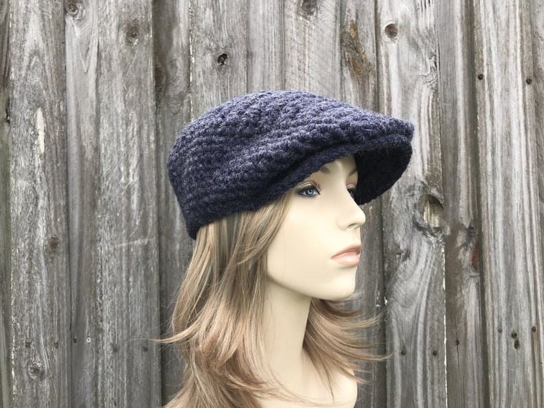 May include: A navy blue newsboy cap made of a textured knit fabric. The cap has a rounded crown and a short, stiff brim. The hat is displayed on a mannequin against a weathered wooden background.