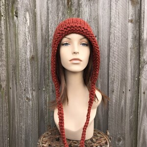 May include: A rust-colored knitted hat with ear flaps and long braided ties. The hat is displayed on a mannequin head against a weathered wooden fence background. The hat's texture and color suggest a handmade, warm accessory.