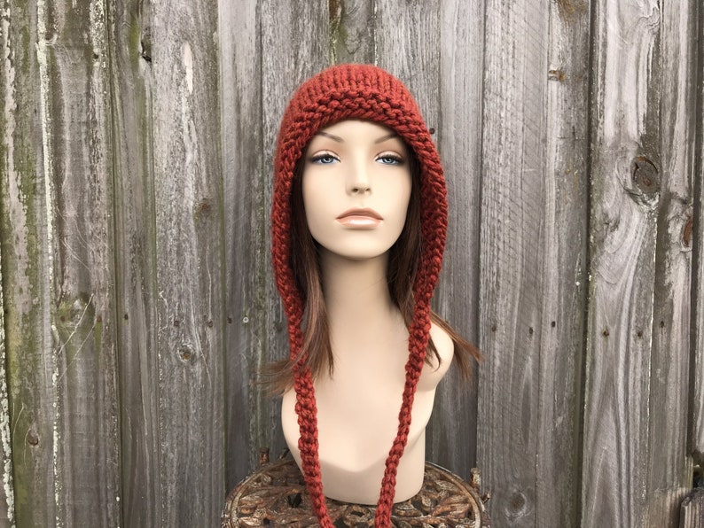 May include: A rust-colored knitted hat with ear flaps and long braided ties. The hat is displayed on a mannequin head against a weathered wooden fence background. The hat's texture and color suggest a handmade, warm accessory.