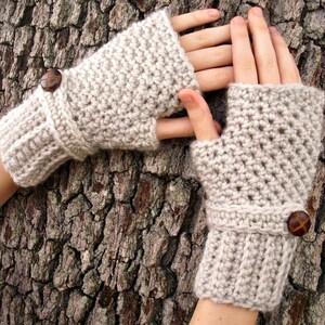 May include: A pair of beige crocheted fingerless gloves with brown buttons. The gloves are shown against a textured bark background.