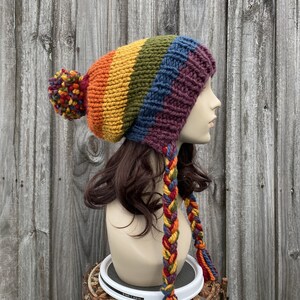 Pu&ograve; includere: Un berretto di lana colorato con un motivo a righe arcobaleno e un pon pon in cima. Il berretto ha lunghi lacci intrecciati che pendono dalla schiena.