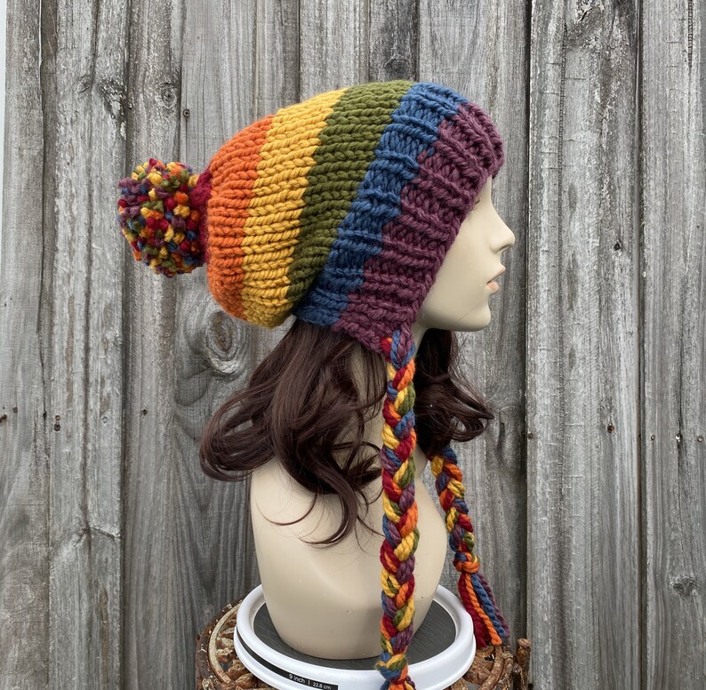 Pu&ograve; includere: Un berretto di lana colorato con un motivo a righe arcobaleno e un pon pon in cima. Il berretto ha lunghi lacci intrecciati che pendono dalla schiena.
