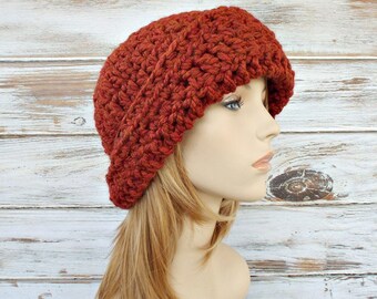 Puede incluir: Un gorro de punto de color naranja óxido oscuro. El gorro tiene un ajuste holgado y está hecho con un punto texturizado.