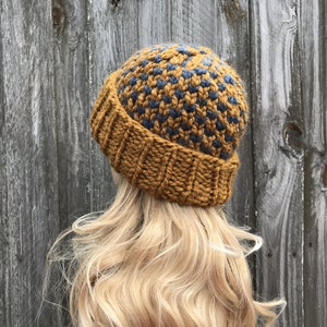 May include: A knitted beanie hat with a mustard yellow and dark blue geometric pattern. The hat is made with a chunky yarn and has a fold-over brim.