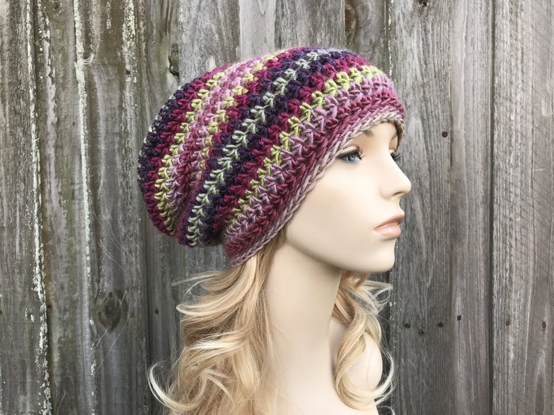 Puede incluir: Un gorro de punto de ganchillo holgado en un patr&oacute;n de rayas multicolores de rosa, morado, verde y marr&oacute;n.