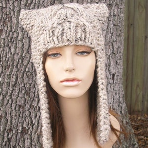 May include: A beige knitted hat with long ear flaps. The hat is made with a cable knit pattern and has a slouchy fit.