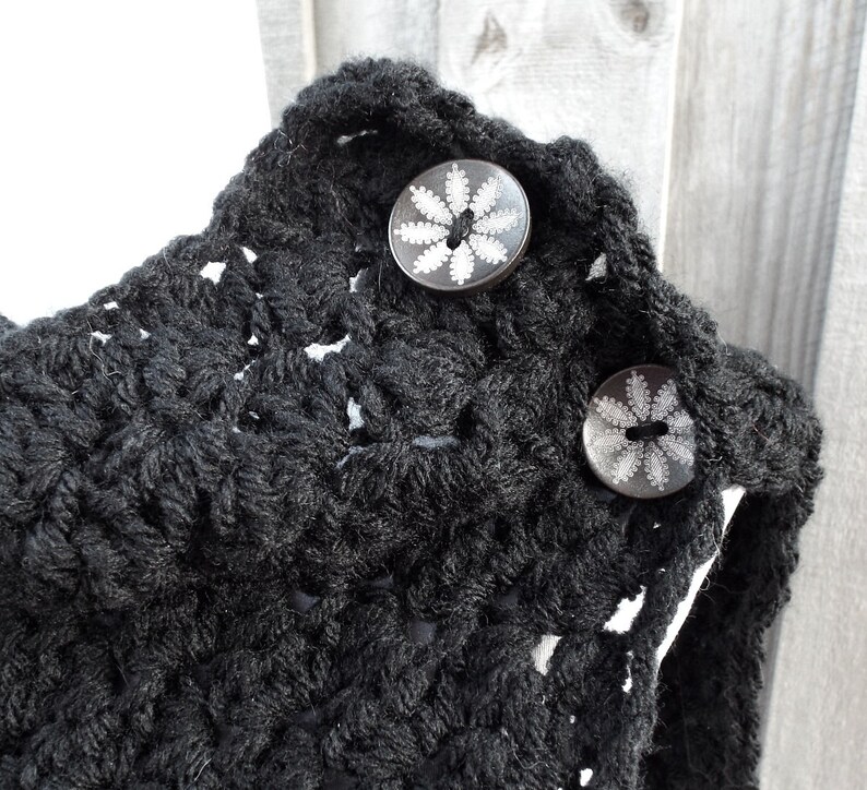 May include: Black crocheted cowl with two decorative buttons featuring a white floral design.