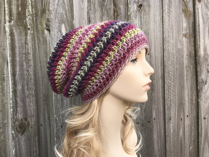Puede incluir: Un gorro de punto de ganchillo holgado en un patr&oacute;n de rayas multicolores de morado, rosa, verde y beige.