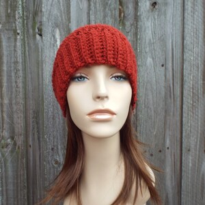 May include: A rust-colored, ribbed knit beanie hat. The hat is displayed on a mannequin head with brown hair and blue eyes. The background is a weathered wooden fence.