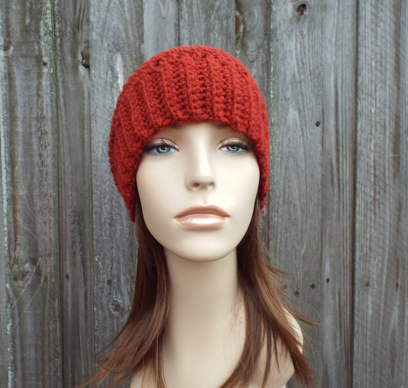 May include: A rust-colored, ribbed knit beanie hat. The hat is displayed on a mannequin head with brown hair and blue eyes. The background is a weathered wooden fence.