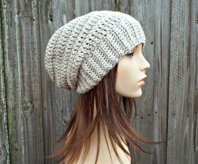 May include: A light beige, slouchy crochet beanie hat. The hat is made of a thick yarn with a ribbed texture. The hat is shown on a mannequin with long brown hair, against a weathered wooden background. The hat is a neutral color.