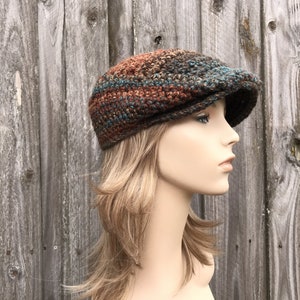 Op de afbeelding: Een bruine, turquoise en oranje gebreide newsboy-pet met een platte bovenkant en een gebogen rand.