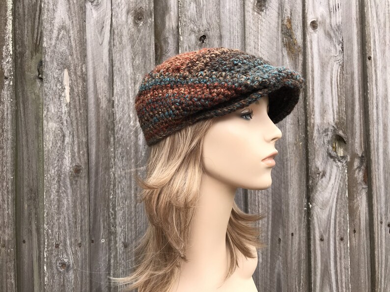 Op de afbeelding: Een bruine, turquoise en oranje gebreide newsboy-pet met een platte bovenkant en een gebogen rand.
