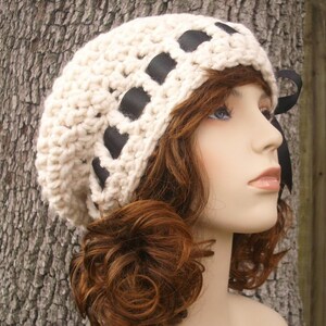 May include: A white crocheted beanie with black ribbon accents. The beanie is worn on a mannequin with brown curly hair.