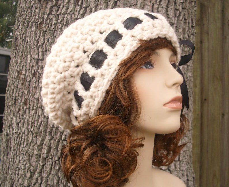May include: A white crocheted beanie with black ribbon accents. The beanie is worn on a mannequin with brown curly hair.