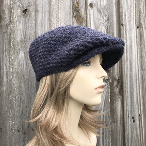 May include: A navy blue crochet newsboy cap with a rounded crown and a short, stiff brim. The hat is made of a textured yarn and is shown against a weathered wooden background. This style of hat is often worn for a casual, vintage look.