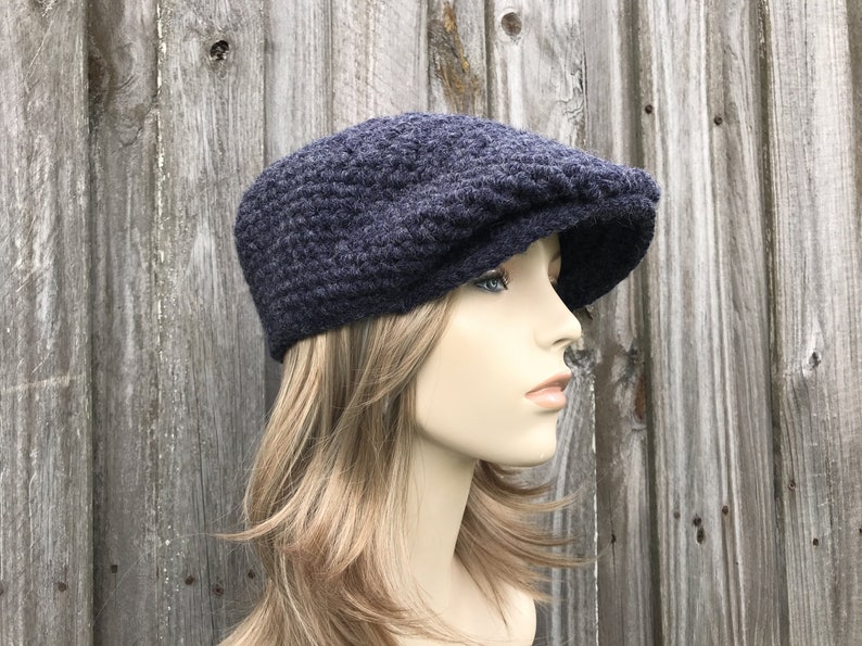 May include: A navy blue crochet newsboy cap with a rounded crown and a short, stiff brim. The hat is made of a textured yarn and is shown against a weathered wooden background. This style of hat is often worn for a casual, vintage look.