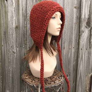 May include: A rust-colored, hand-knitted hat with ear flaps and long braided ties. The hat is displayed on a mannequin head, showcasing its texture and design. The hat is made of thick yarn, perfect for cold weather.