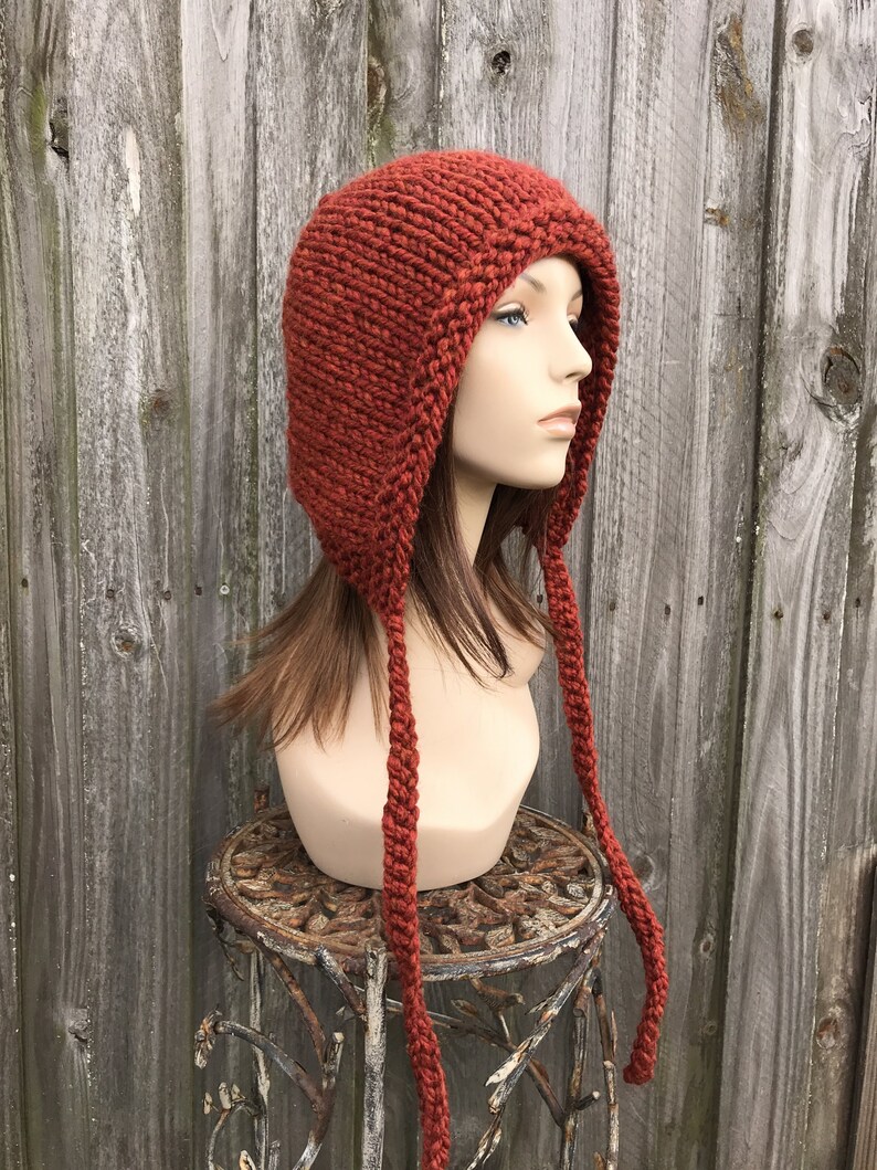 May include: A rust-colored, hand-knitted hat with ear flaps and long braided ties. The hat is displayed on a mannequin head, showcasing its texture and design. The hat is made of thick yarn, perfect for cold weather.