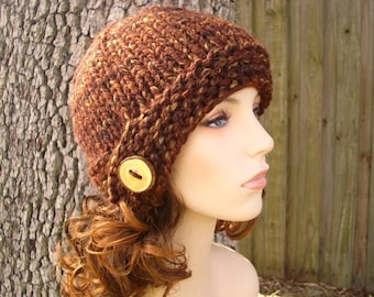 Puede incluir: Un gorro de punto marrón y dorado con un botón en el lateral. El gorro está hecho con un hilo grueso y tiene un ajuste holgado.