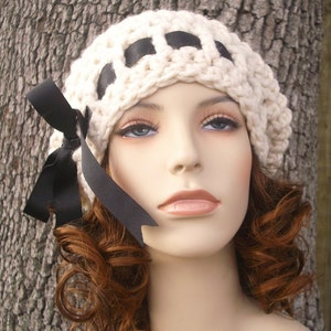 May include: A white crocheted beanie with black ribbon accents and a black bow. The beanie is worn on a mannequin with curly brown hair.