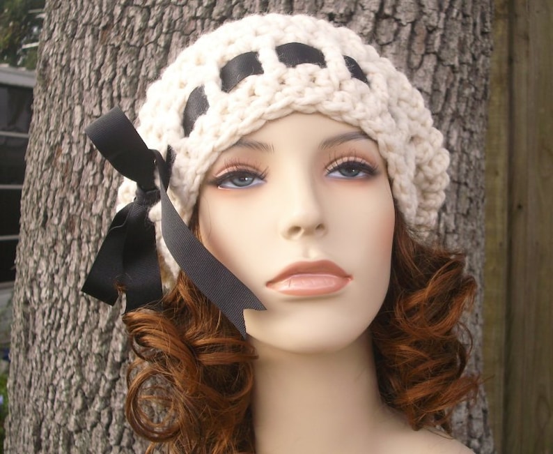 May include: A white crocheted beanie with black ribbon accents and a black bow. The beanie is worn on a mannequin with curly brown hair.