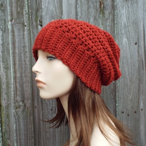 May include: A rust-colored, handmade crochet beanie hat. The hat is made with a ribbed stitch pattern, giving it a textured appearance. The hat is shown on a mannequin head, with a wooden background.