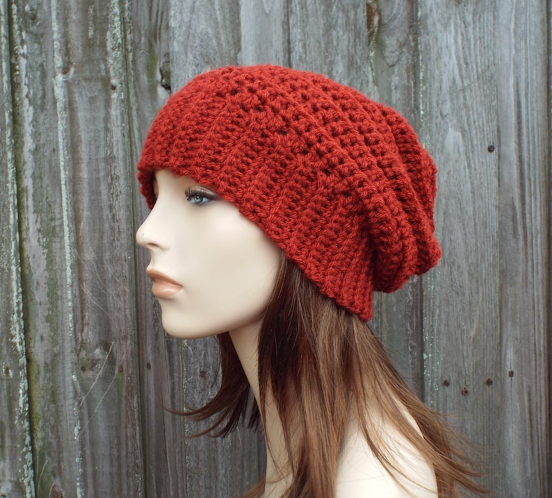 May include: A rust-colored, handmade crochet beanie hat. The hat is made with a ribbed stitch pattern, giving it a textured appearance. The hat is shown on a mannequin head, with a wooden background.