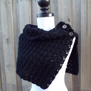 May include: Black crocheted shawl with a triangular shape and two decorative buttons. The shawl is displayed on a white mannequin against a wood background.