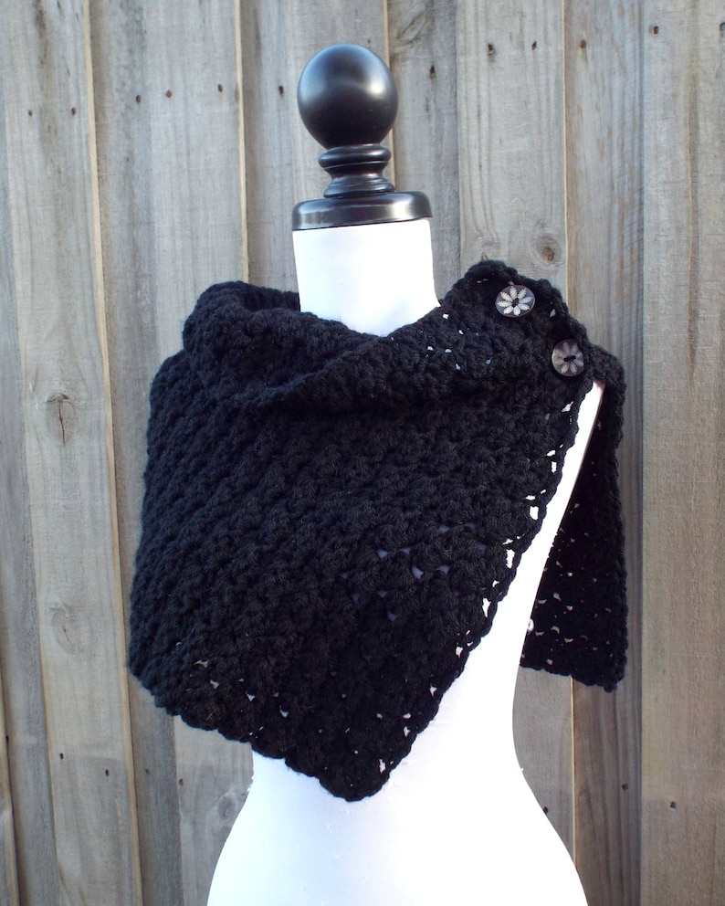 May include: Black crocheted shawl with a triangular shape and two decorative buttons. The shawl is displayed on a white mannequin against a wood background.
