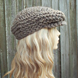 May include: A brown newsboy cap crafted from thick, chunky knit yarn. The hat features a textured, multi-tonal brown appearance with a prominent brim extending from the front. The knit construction displays a visible, repeating stitch pattern.