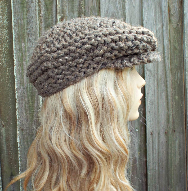 May include: A brown newsboy cap crafted from thick, chunky knit yarn. The hat features a textured, multi-tonal brown appearance with a prominent brim extending from the front. The knit construction displays a visible, repeating stitch pattern.