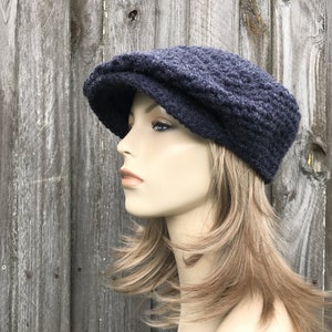 May include: A dark blue, knitted newsboy cap with a textured pattern. The cap has a rounded crown and a small, stiff brim. The hat is displayed on a mannequin with blonde hair against a weathered wood background.