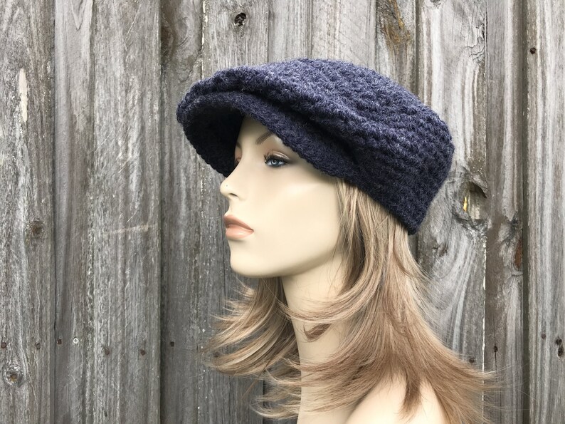 May include: A dark blue, knitted newsboy cap with a textured pattern. The cap has a rounded crown and a small, stiff brim. The hat is displayed on a mannequin with blonde hair against a weathered wood background.
