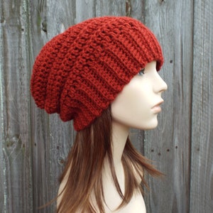 May include: A rust-colored, slouchy beanie hat made of a thick, textured knit. The hat is displayed on a mannequin head, showcasing its ribbed design and relaxed fit. The warm, earthy tone of the hat suggests a cozy, winter accessory.