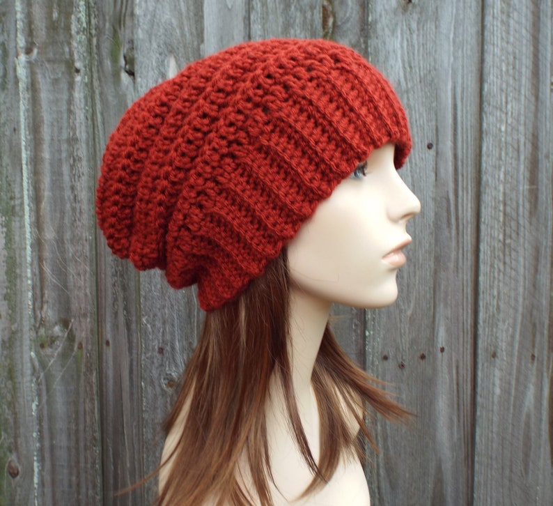 May include: A rust-colored, slouchy beanie hat made of a thick, textured knit. The hat is displayed on a mannequin head, showcasing its ribbed design and relaxed fit. The warm, earthy tone of the hat suggests a cozy, winter accessory.