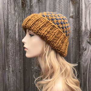 May include: A mustard yellow and blue knitted beanie hat with a textured pattern. The hat is worn on a mannequin's head with long blonde hair.