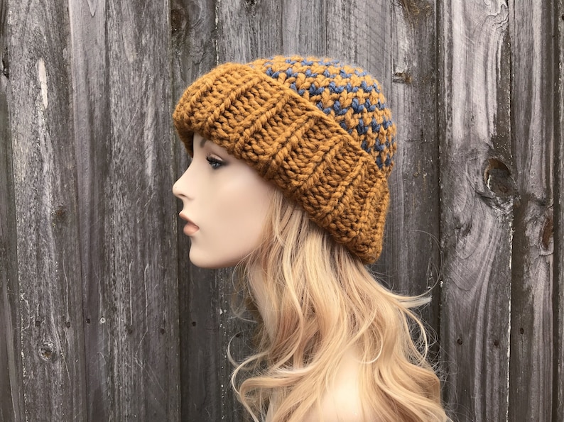 May include: A mustard yellow and blue knitted beanie hat with a textured pattern. The hat is worn on a mannequin's head with long blonde hair.