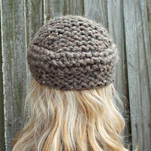 May include: A brown knit beanie with a thick, textured garter stitch pattern. The yarn features subtle flecks of lighter tan and dark gray throughout the material, creating a heathered appearance. The hat has a rounded crown and a folded brim.