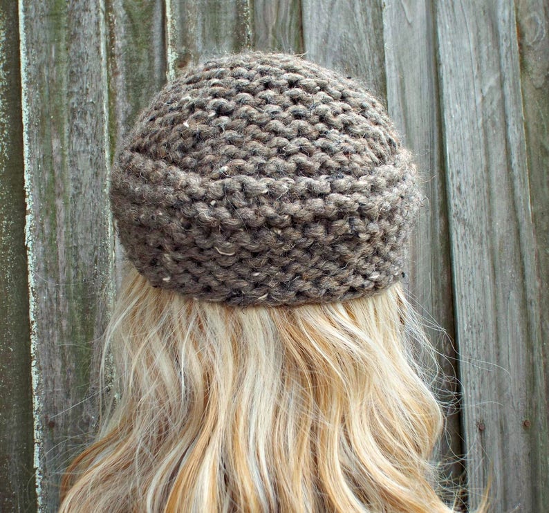 May include: A brown knit beanie with a thick, textured garter stitch pattern. The yarn features subtle flecks of lighter tan and dark gray throughout the material, creating a heathered appearance. The hat has a rounded crown and a folded brim.