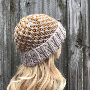 May include: A knitted beanie hat in a light grey and mustard yellow color combination. The hat has a ribbed brim and a textured pattern.