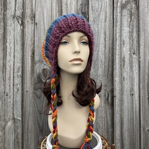 Pu&ograve; includere: Un cappello di lana con lunghe trecce colorate. Il cappello &egrave; fatto di un filato morbido e spesso in tonalit&agrave; di viola, blu, arancione e verde. Il cappello &egrave; indossato da un manichino con i capelli castani.