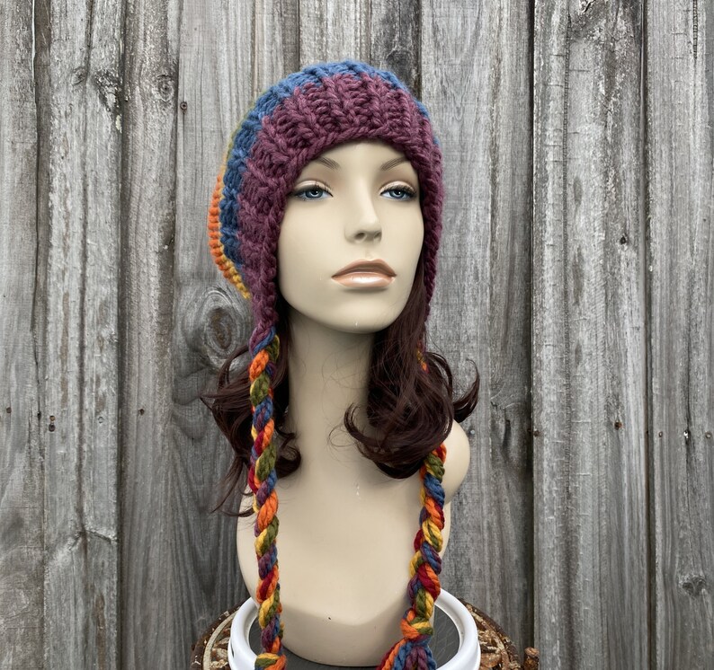 Pu&ograve; includere: Un cappello di lana con lunghe trecce colorate. Il cappello &egrave; fatto di un filato morbido e spesso in tonalit&agrave; di viola, blu, arancione e verde. Il cappello &egrave; indossato da un manichino con i capelli castani.