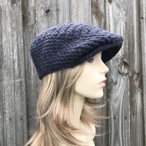 May include: A navy blue, crocheted newsboy cap with a textured pattern. The cap has a rounded crown and a short, stiff brim. The hat is displayed on a mannequin head against a weathered wooden background.