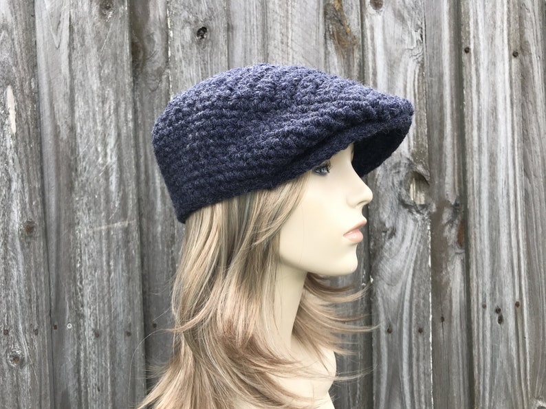 May include: A navy blue, crocheted newsboy cap with a textured pattern. The cap has a rounded crown and a short, stiff brim. The hat is displayed on a mannequin head against a weathered wooden background.