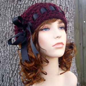 May include: A burgundy crocheted beanie with a black ribbon bow. The beanie has a wide band of black ribbon woven into the fabric.