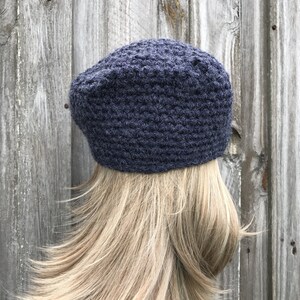 May include: A dark blue crocheted beret hat. The hat is made of a textured yarn and has a rounded crown. The beret is worn on a person with blonde hair, against a weathered wooden background.